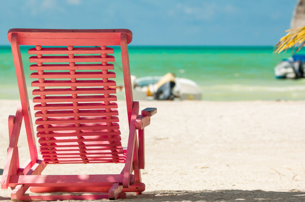 A bright pink wooden lounge chair facing the turquoise ocean on a sandy beach. A small boat is visible in the distance, and the sky is clear with a few clouds.