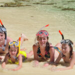 A smiling family wearing snorkeling gear, lying on the shore of a clear, shallow beach with coral rocks in the background.