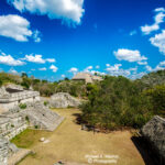 ncient Maya ruins with stone pyramids and structures under a bright blue sky, surrounded by lush jungle.