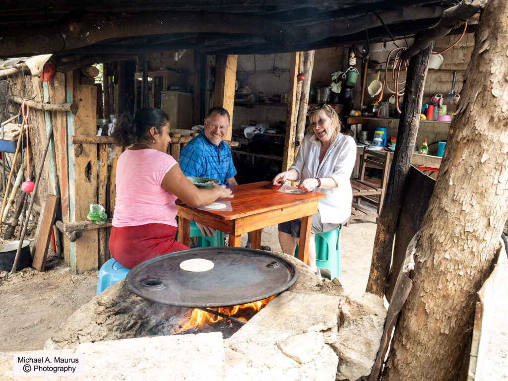 Three people sitting at a small wooden table in an open-air rustic kitchen, smiling and preparing tortillas together. A fire is burning in an open stone oven with tortillas cooking on a metal pan.