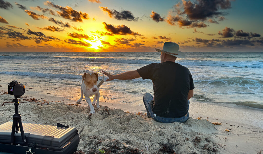 A man with a wide-brimmed hat sitting on the beach at sunrise, reaching out to a dog, with a camera on a tripod nearby and waves in the background.