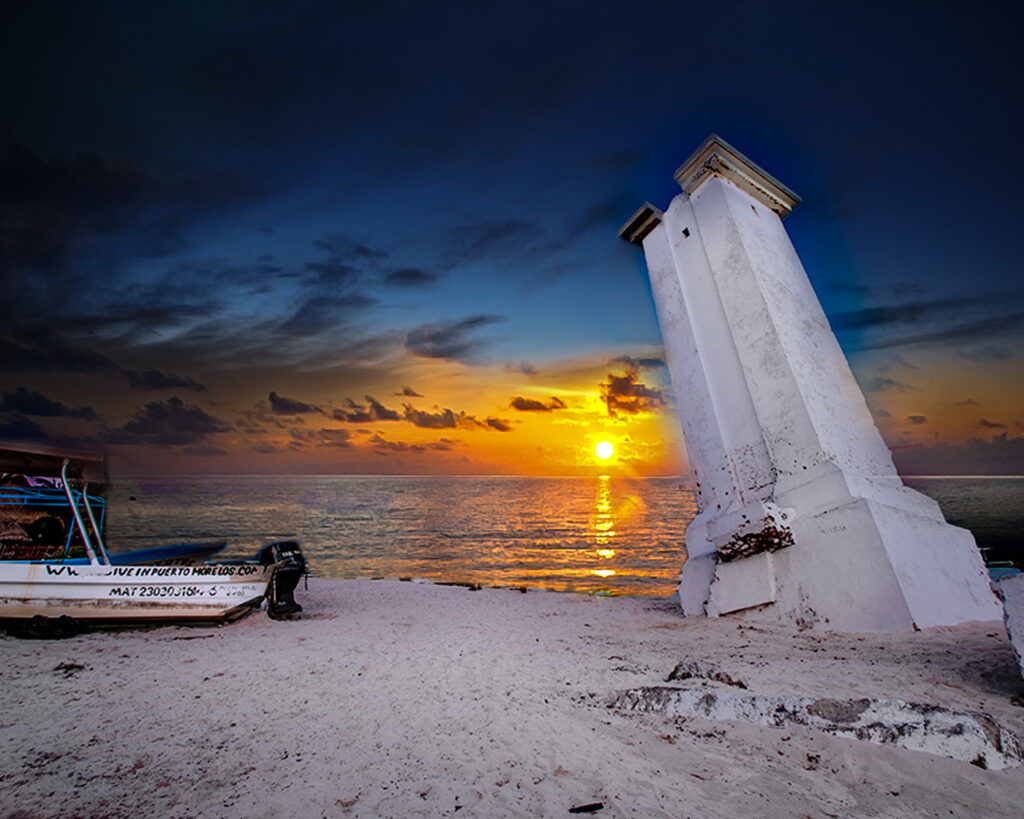 A sunrise over Puerto Morelos with a white lighthouse on the left, vibrant sky colors, and boats floating on the calm sea.