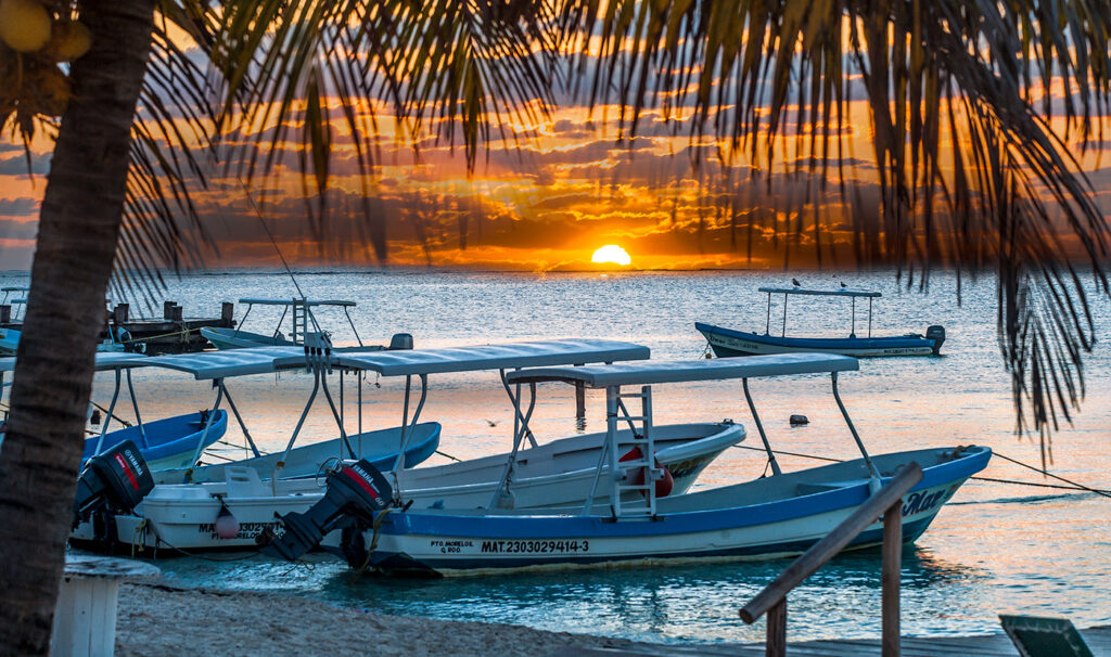 Boats resting in shallow waters at sunrise, framed by palm fronds, with a sky filled with warm colors reflecting on the water.