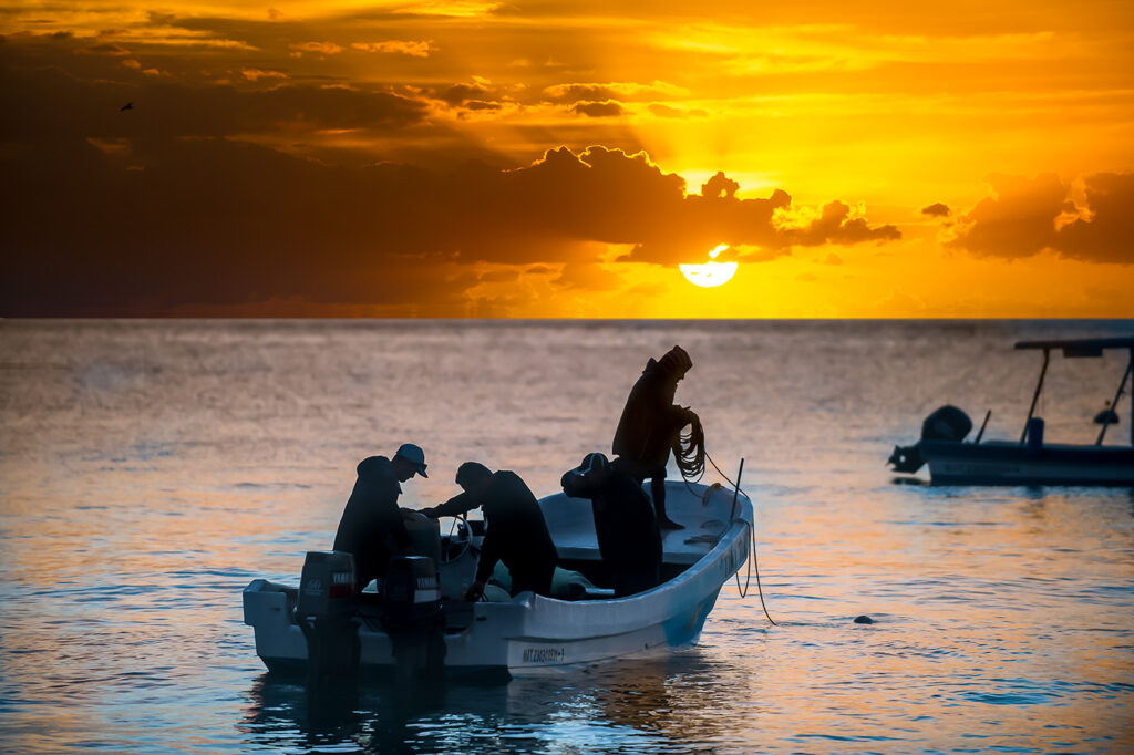 Silhouetted fishermen preparing their boat at sunrise, with a golden sky and calm sea in the background.