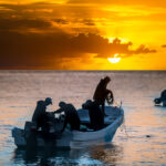 Fishermen in a small boat on the ocean at sunrise, silhouetted against a golden sky.