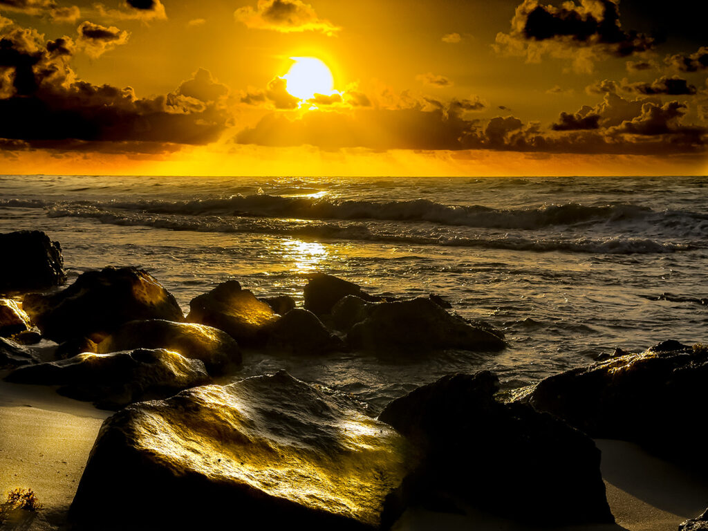 The sun rising over the ocean, illuminating the rocky shore with golden light, and dramatic clouds enhancing the scene.