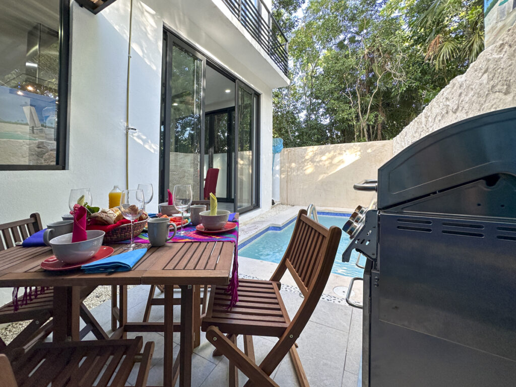 An outdoor dining area beside a small pool, with a grill and wooden patio furniture set up for a meal.