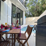 An outdoor dining area beside a small pool, with a grill and wooden patio furniture set up for a meal.