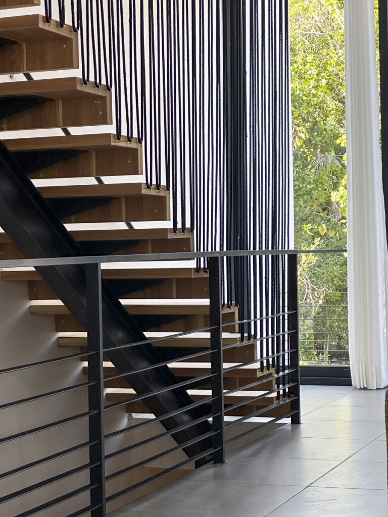 A modern staircase with minimalist wooden steps and sleek black railings, set against a backdrop of large windows that reveal a lush, green forest outside. Light flows into the space, highlighting the clean lines and contemporary design.