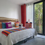 A bright bedroom with a large wooden headboard, colorful pillows, and floor-to-ceiling windows overlooking the trees.