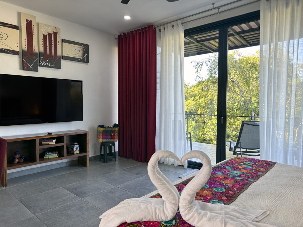 A bedroom with heart-shaped towel swans on the bed, a large TV, and sliding doors leading to a balcony.