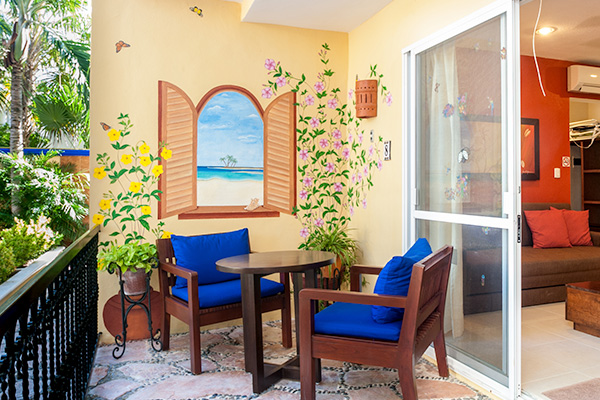 A small balcony with two wooden chairs and a table, featuring a colorful wall mural of a beach view.