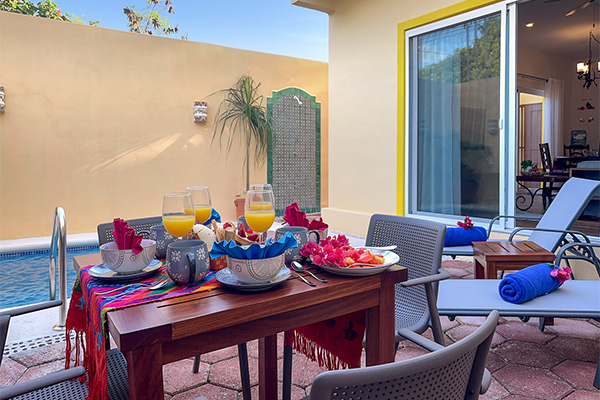 An outdoor patio with a table set for breakfast beside a small pool, with lounge chairs and towels nearby.