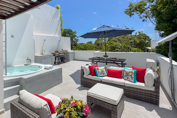 A rooftop terrace with a hot tub, lounge seating, an outdoor dining area, and a grill, all shaded by a large umbrella.