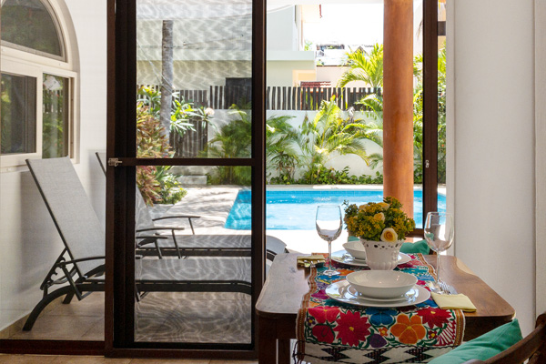 View of a pool and tropical courtyard from inside a dining area, with a table set for two and lounge chairs visible outside.