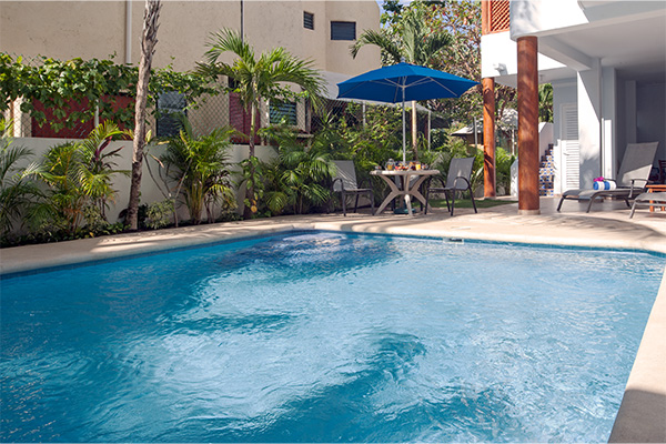 A clear blue swimming pool in a tropical courtyard with a shaded seating area under a blue umbrella.