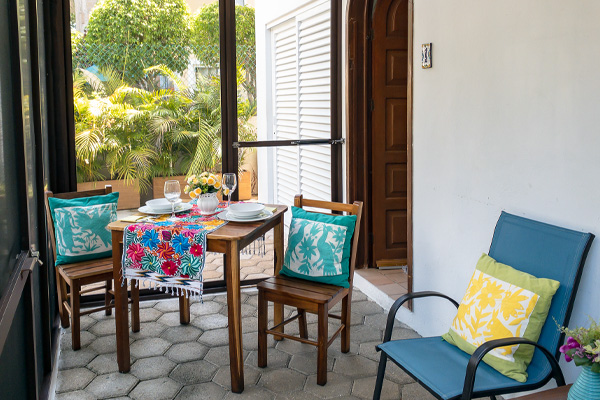 A cozy outdoor patio with a wooden dining table set for two, featuring colorful cushions and a decorative tablecloth. The entrance to the Sonoma Apartment 1 is visible, with a wooden door and white shutters, surrounded by lush greenery.