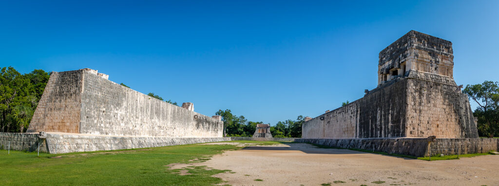 A wide panoramic view of the Great Ball Court at Chichen Itza, with towering stone walls on either side and stone hoops visible high up on the walls. The court stretches into the distance under a clear blue sky, surrounded by greenery and trees at the far end, highlighting the expansive and impressive scale of the ancient structure.