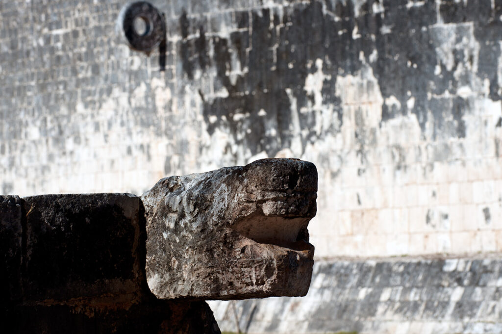 A close-up of a stone serpent head sculpture at the Great Ball Court in Chichen Itza, with the wall and one of the stone rings visible in the background. The serpent head is weathered but well-defined, adding to the mysterious atmosphere of this ancient Maya sporting arena.