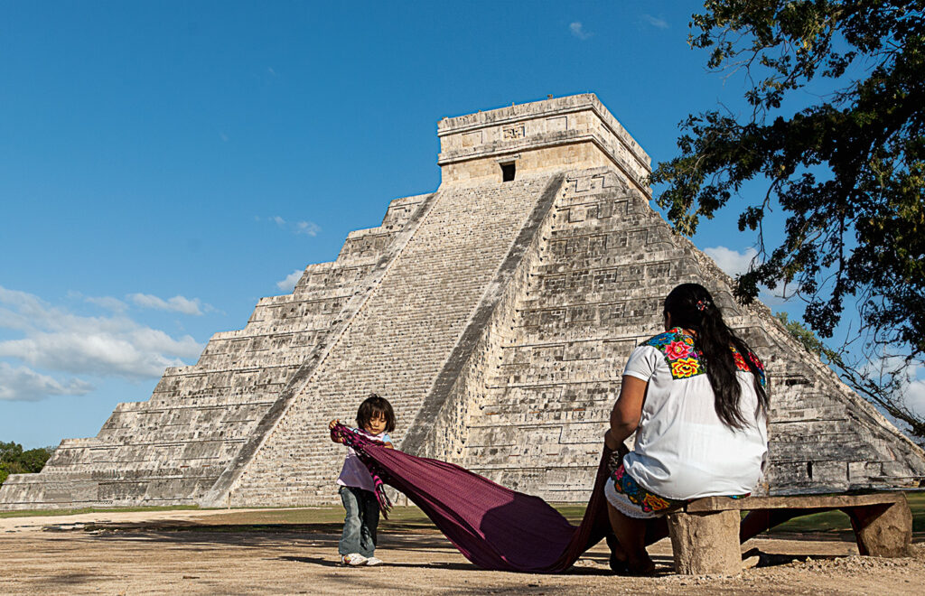 A woman dressed in traditional Maya clothing sits on a bench near the Kukulkan Pyramid at Chichen Itza while a young child plays with a colorful cloth in front of her. The iconic pyramid rises in the background against a clear blue sky.