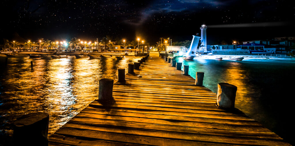 A wooden dock at night, illuminated by golden lights, leading toward the iconic leaning lighthouse in Puerto Morelos under a starry sky.