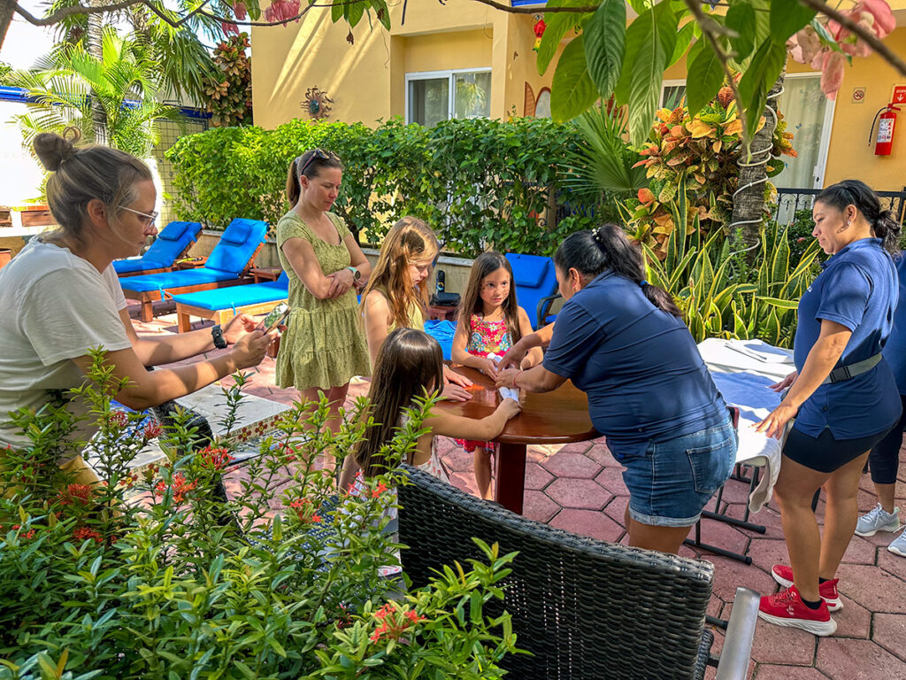  Housekeeping staff at Abbey del Sol demonstrate towel folding techniques to a group of young guests, who watch intently and participate in creating towel art. Families observe the interaction, capturing the moment by the poolside in a lush garden setting.