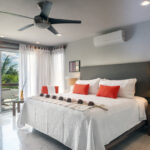 A bright, modern bedroom with a king-sized bed, white linens, and red accent pillows, overlooking a private balcony with lush palm trees.