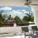 A private balcony with modern seating, ceiling fan, and views of lush palm trees and the ocean in the distance.