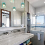 A modern bathroom with a double vanity, turquoise pendant lights, a walk-in shower, and a large mirror reflecting natural light.