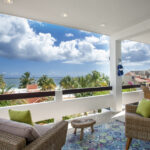 A spacious terrace with wicker furniture, colorful cushions, and a vibrant rug, overlooking palm trees and the ocean under a bright blue sky.