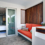 A cozy corner on a balcony with a built-in orange cushioned bench, colorful pillows, and wood panel accents, leading into a bedroom.