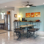 A dining area with a glass-top table, wrought iron chairs, colorful wall art, and a modern kitchen in the background.