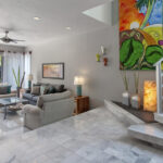 A bright living room with modern sofas, vibrant artwork, marble flooring, and a staircase leading to the upper level.