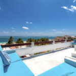 A rooftop terrace with bright blue seating, overlooking the ocean and palm trees under a clear blue sky.