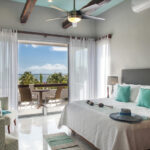 A bright and airy bedroom with a king-sized bed, turquoise accents, a sitting area, and a balcony overlooking the ocean and palm trees.