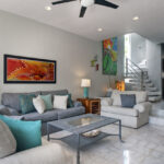 A spacious living room with gray and cream sofas, turquoise accent pillows, vibrant artwork, and a staircase leading to the upper floor.