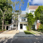 A modern three-story home surrounded by lush greenery, with clean lines, a spacious driveway, and a rooftop terrace peeking above the treetops.