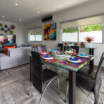 A dining area with colorful table settings and a cozy living room in the background, decorated with vibrant Mexican art and lush greenery visible through large windows.