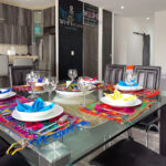 A close-up view of a glass dining table set with colorful embroidered placemats, napkins folded into intricate shapes, and sparkling glassware.