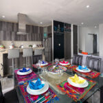 A stylish, open-concept dining area with a glass table set with colorful napkins, leading into a modern kitchen with sleek cabinetry and a cozy living space in the background.