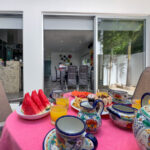 A colorful breakfast table set with fresh fruit, traditional Mexican pottery, and glasses of orange juice, overlooking a modern open-concept kitchen and dining area through sliding glass doors.