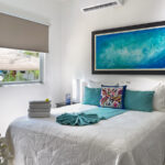 A bright and airy bedroom featuring a double bed with turquoise accents, a colorful throw pillow, and a large ocean-inspired painting above the bed.