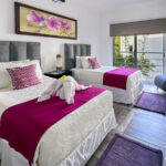 A bedroom with two twin beds dressed in magenta accents, a vibrant orchid painting on the wall, and large windows letting in natural light.