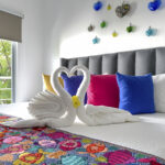 A close-up of swan-shaped towels resting on a bed with colorful pillows and a detailed embroidered blanket, against a backdrop of glass heart decorations.