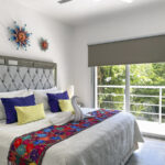 A bedroom with a king-size bed, decorative sunburst wall art, colorful throw pillows, and a balcony overlooking lush greenery.