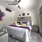 A serene bedroom featuring a king-size bed with vibrant throw pillows, a colorful embroidered blanket, decorative sunburst wall art, and floor-to-ceiling sliding glass doors leading to a balcony.