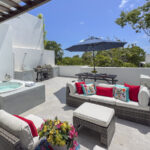 A spacious rooftop terrace with a wicker seating area, colorful pillows, an umbrella-shaded dining table, and a BBQ kitchen setup, surrounded by greenery.