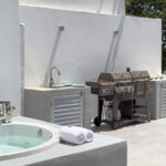 An outdoor kitchen area with a stainless-steel BBQ, a sink, and a Jacuzzi, set against minimalist white walls and surrounded by lush greenery.