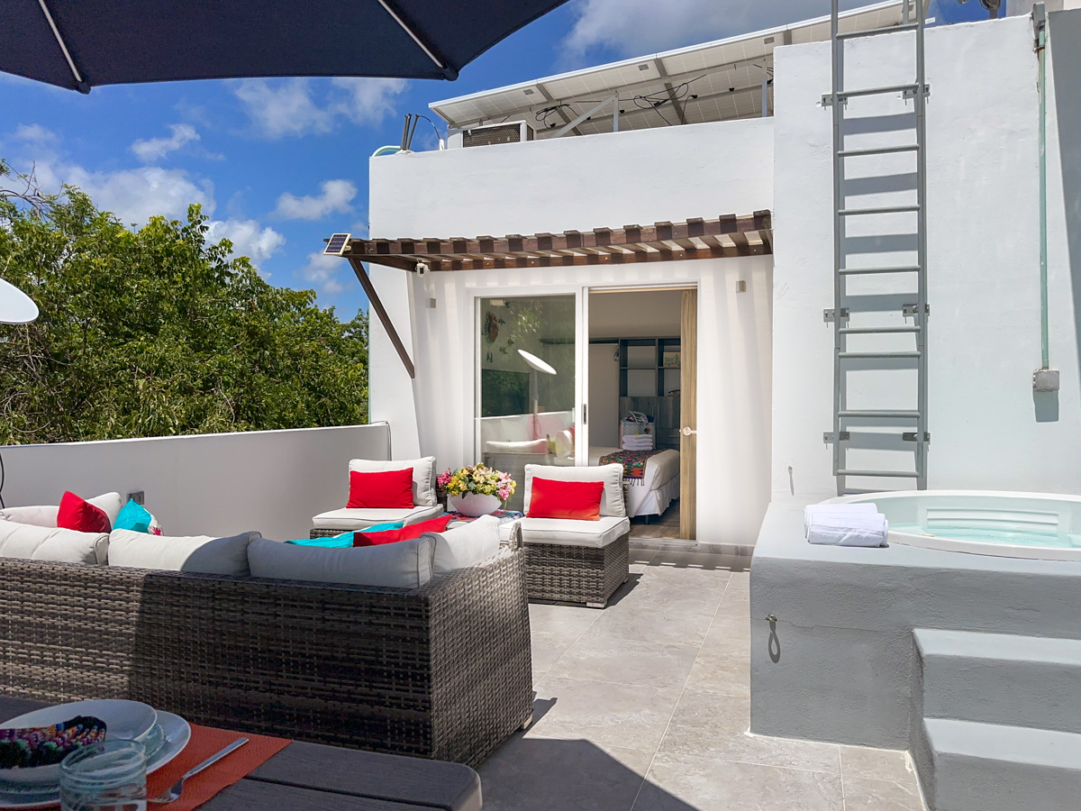 A modern rooftop terrace featuring a Jacuzzi, comfortable seating with bright red and turquoise cushions, and sliding doors leading to an indoor suite, surrounded by lush greenery under a clear blue sky.