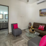 A cozy living room with a gray sofa, bright pink cushions, and a glass coffee table, located near a bathroom with double sinks.